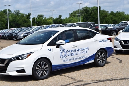 a white Nissan Versa with a blue NMMC Home Health and Hospice logo. The female driver is waving and smiling through the window.