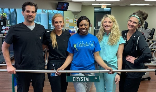 Crystal Lane poses with her rehab team in front of the Crystal Ln street sign on the parallel bars