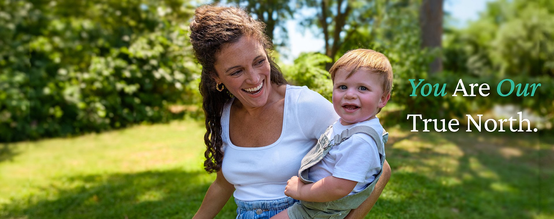 A mother in a white top and jeans with curly brown hair smiles at her son on her hip. He is wearing khaki overalls and a white shirt and looking toward the camera. Text on the image says You Are Our True North.