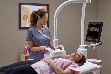 a woman in a pink hospital gown lies on her back on a table while a technician administers an ultrasound of her right breast